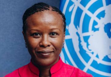 A woman wearing a pink blouse stands in front of a blue background and behind her is the UN flag 