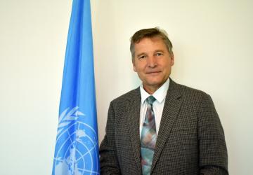 The Resident Coordinator stands in front of a United Nations flag and is wearing a brown suite