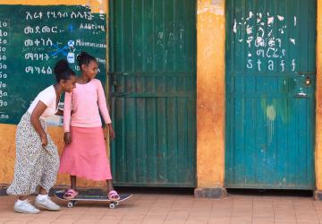 Two girls in front of a classroom, one of them is on a skateboard.