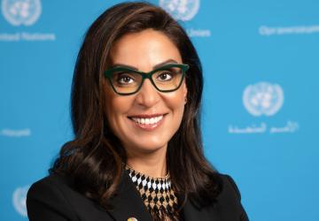 The RC is looking at the camera and smiling. She is wearing a black blazer and glasses. Behind her is a backdrop with the UN logo
