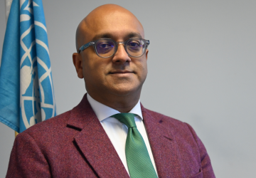 The RC is standing in front of a grey background with the UN flag behind him. He is wearing a red jacket and glasses