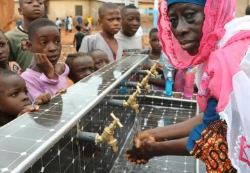 A group of children and a woman gather around water faucets with running water