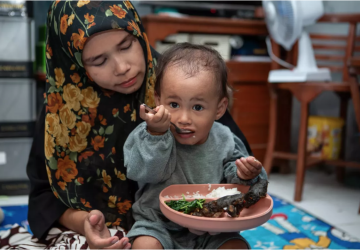 A little boy is eating his food seated on his mother's lap