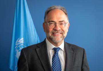 The RC is looking at the camera and smiling wearing a blue tie and black suit with the UN Flag