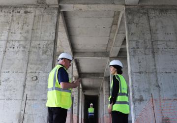 Two people are standing in front of a large infrastructure project speaking to each other 