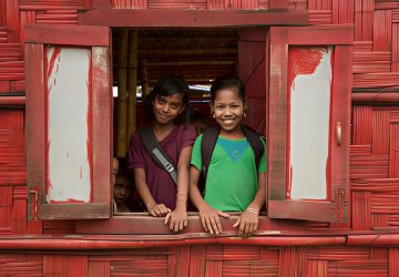 Two girls smile while standing in a window frame of a red house.
