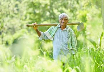 A smiling farmer carries a hoe on his shoulders while standing in a green field.