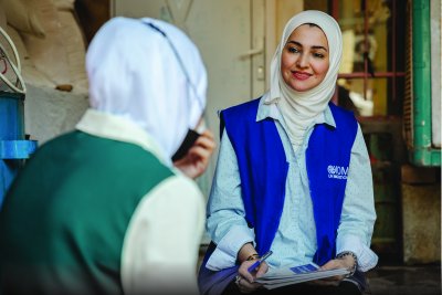 A person in a blue vest with an IOM logo writes notes while speaking with another person wearing a headscarf in a modest indoor setting.