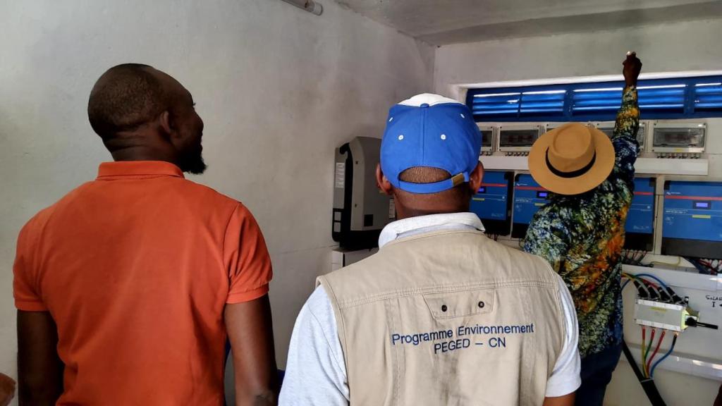 A group of people stand in front of a switch board. One of them is wearing a vest that says UNDP Guinea (in French)