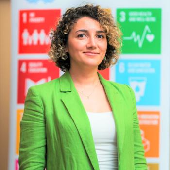 A woman in a green jacket stands in front of the SDG sign.