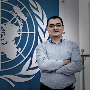 A man in a blue shirt stands in front of the United Nations logo.