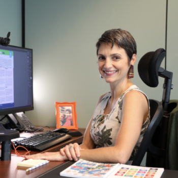 A woman smiles at the camera from her desk. 