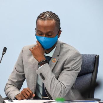 A man in a suit and a blue mask sit at a table with a microphone.
