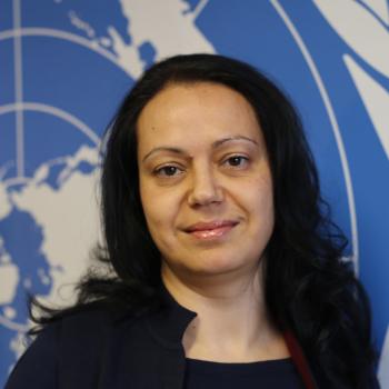 A woman with black hair stands in front of the United Nations logo.