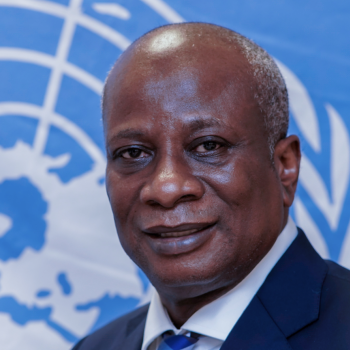A man with a kind face smiles at the camera in front of the United Nations logo.