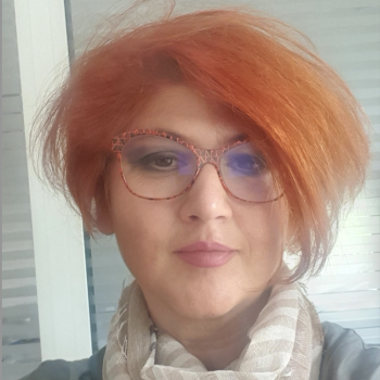 A woman with red hair and peach glasses looks directly at the camera.