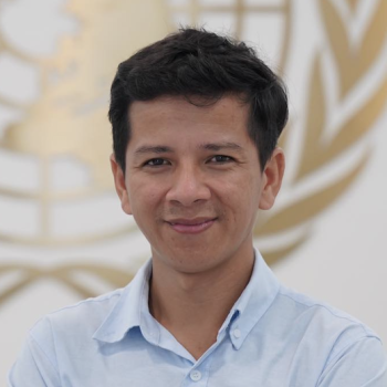 A man, with a kind face, smiles directly at the camera in front of the United Nations logo.