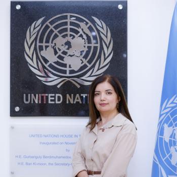 A woman wearing a beige dress, standing next to the UN flag.