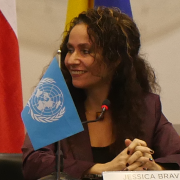 A woman sits at a meeting with a flag. 