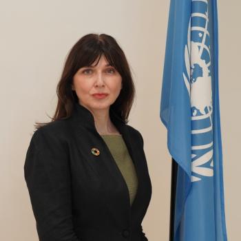 A woman in a black suit stands next to the blue United Nations flag.