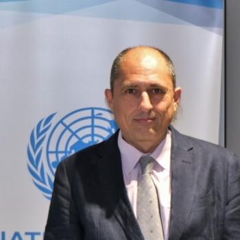 A man in a suit and tie stands in front of a blue and white United Nations logo.