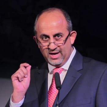 A man in a pink tie and blue suit speaks into a microphone.