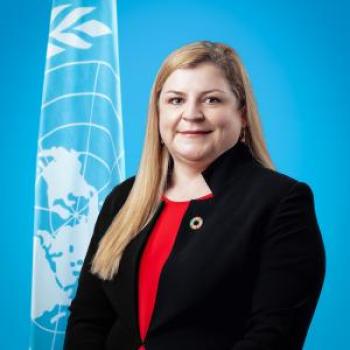 A woman with a black suit and red shirt stands next to the United Nations Flag.