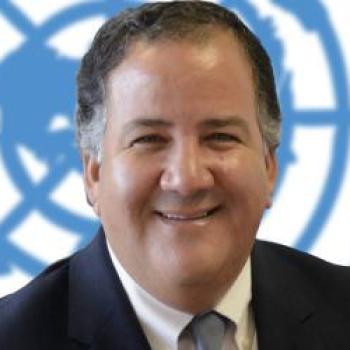 A smiling man stands in front of the United Nations logo.