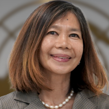 A woman in a pearl necklace smiles directly at the camera in front of a United Nations Logo.