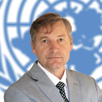 A man in a grey suit looks directly at the camera with the United Nations behind him.