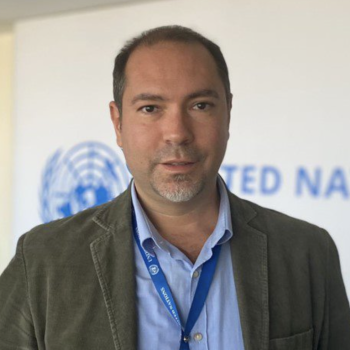 A man with a blue shirt and green jacket looks directly at the camera in front of the United Nations logo.