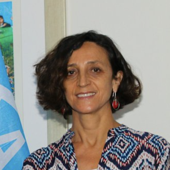 A woman with brown hair, wears a colourful shirt and smiles directly at the camersa.