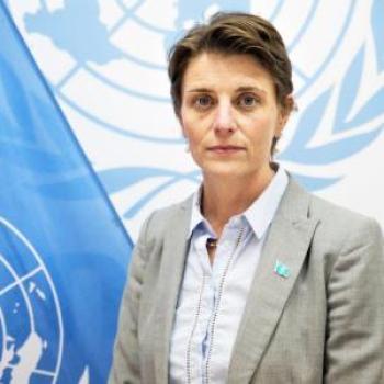 A woman in a grey suit and white shirt stands near the UN logo and flag.