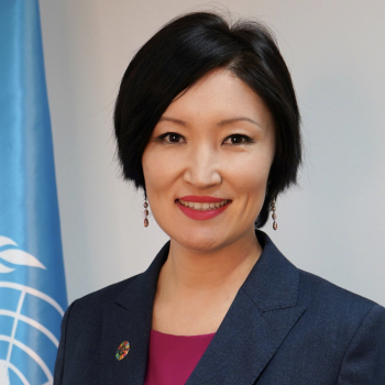 A woman smiles directly at the camera next to a United Nations flag.