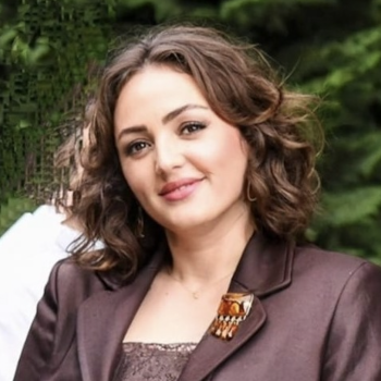 A woman in a brown suit stands outside near some trees.