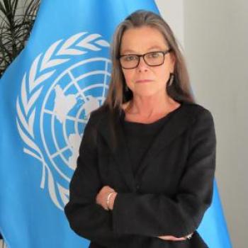 A woman with glasses stands in front of the United Nations flag with her arms crossed.