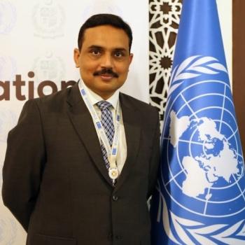 man in dark suit stands next to UN flag 