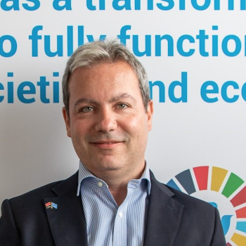 A man in a suit, with UN logos and phrases behind him, smiles at the camera.