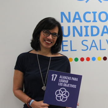 women in dark top stands in front of white background holding a small sign 