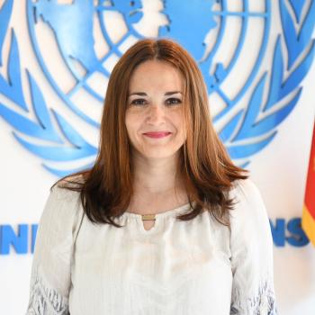 woman in white top stands in front of UN emblem 