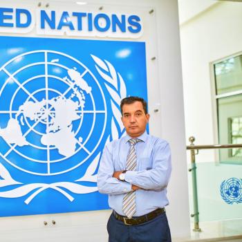 man with his arms crossed stands in front of UN flag 