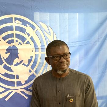 man in brown tunic stands in front of UN flag 