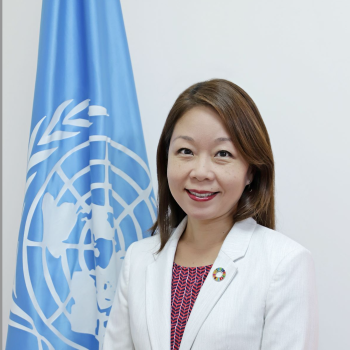 woman in white suit stands next to UN flag 