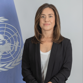woman in dark suit stands in front of UN flag 