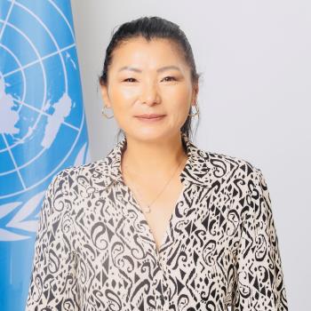 woman in patterned shirt and dark hair stands in front of UN flag