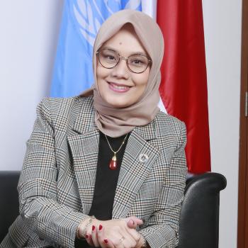 woman in grey blazer and light headscarf smiles to the camera with UN flag behind