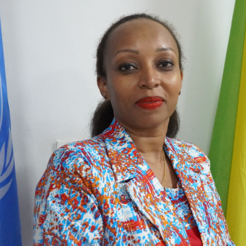 A woman in a colourful jacket standing in front of the UN flag and another flag by her side