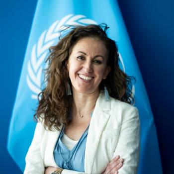 A woman looks at the camera and smiles, the UN flag is bhind her and she is wearing a white jacket