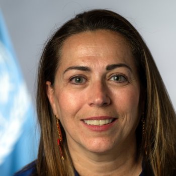 A woman is looking at the camera and smiling with the UN flag behind her