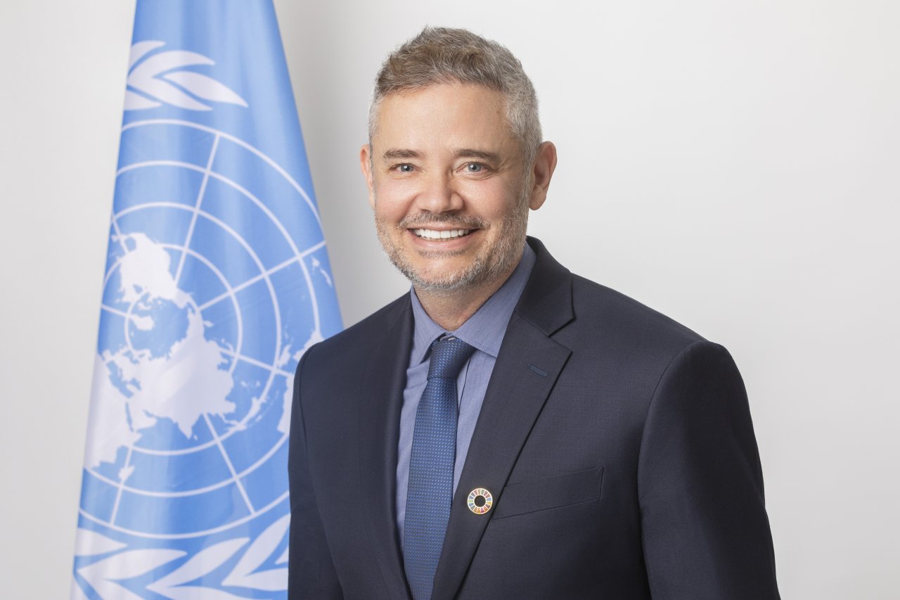 man in dark suit stands next to UN flag 